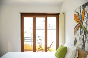 Un dormitori amb un llit, un coixí verd, un capçal de fusta, un quadre i una porta corredissa de vidre que dona a una zona exterior amb una taula i cadires.