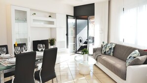 A modern living room with a glass dining table, black chairs, a grey sofa with patterned cushions, and a glass coffee table.