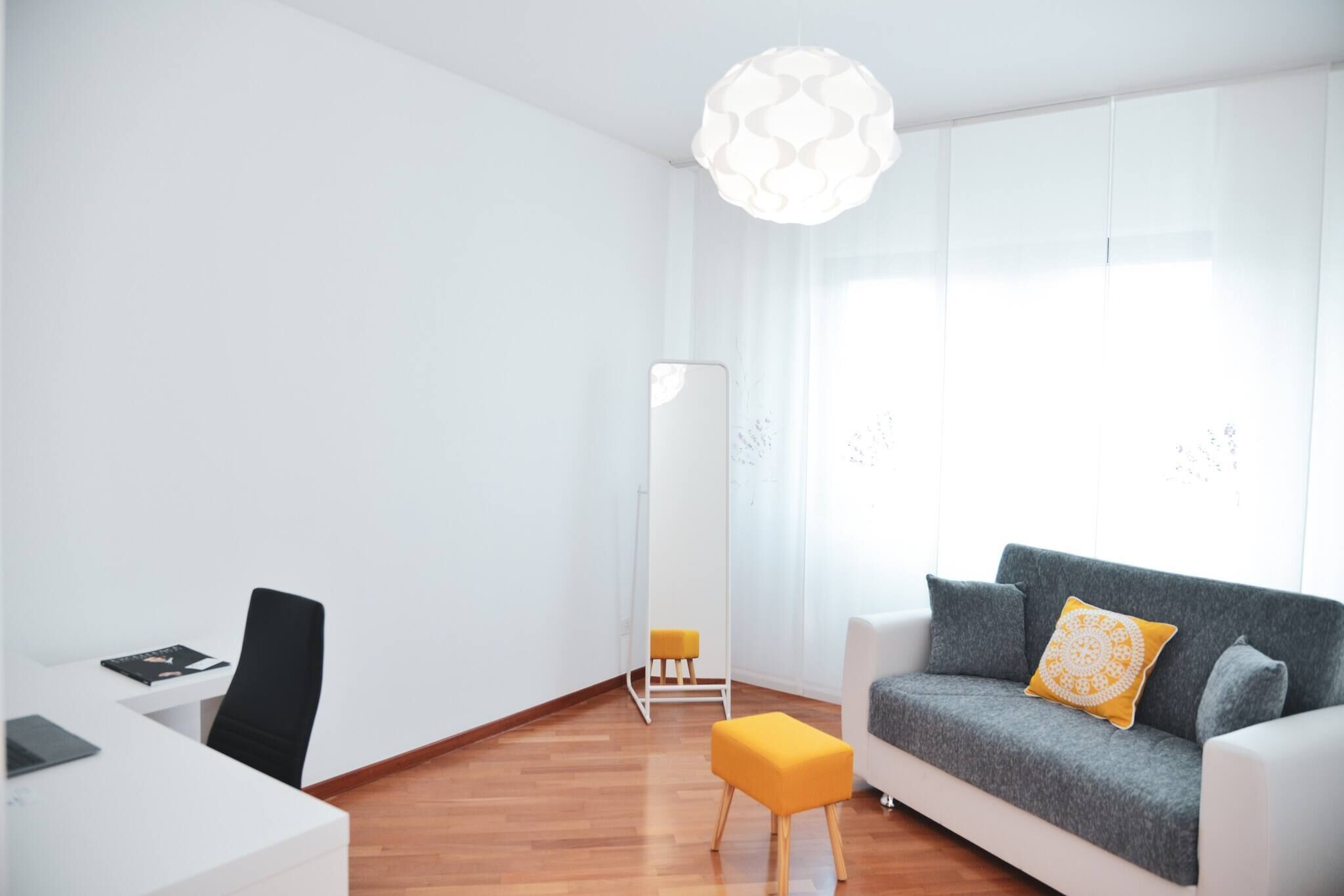 Ein modernes Wohnzimmer mit grauem Sofa, weißem Schreibtisch, schwarzem Sessel, einem gelben Hocker und einem großen Spiegel.