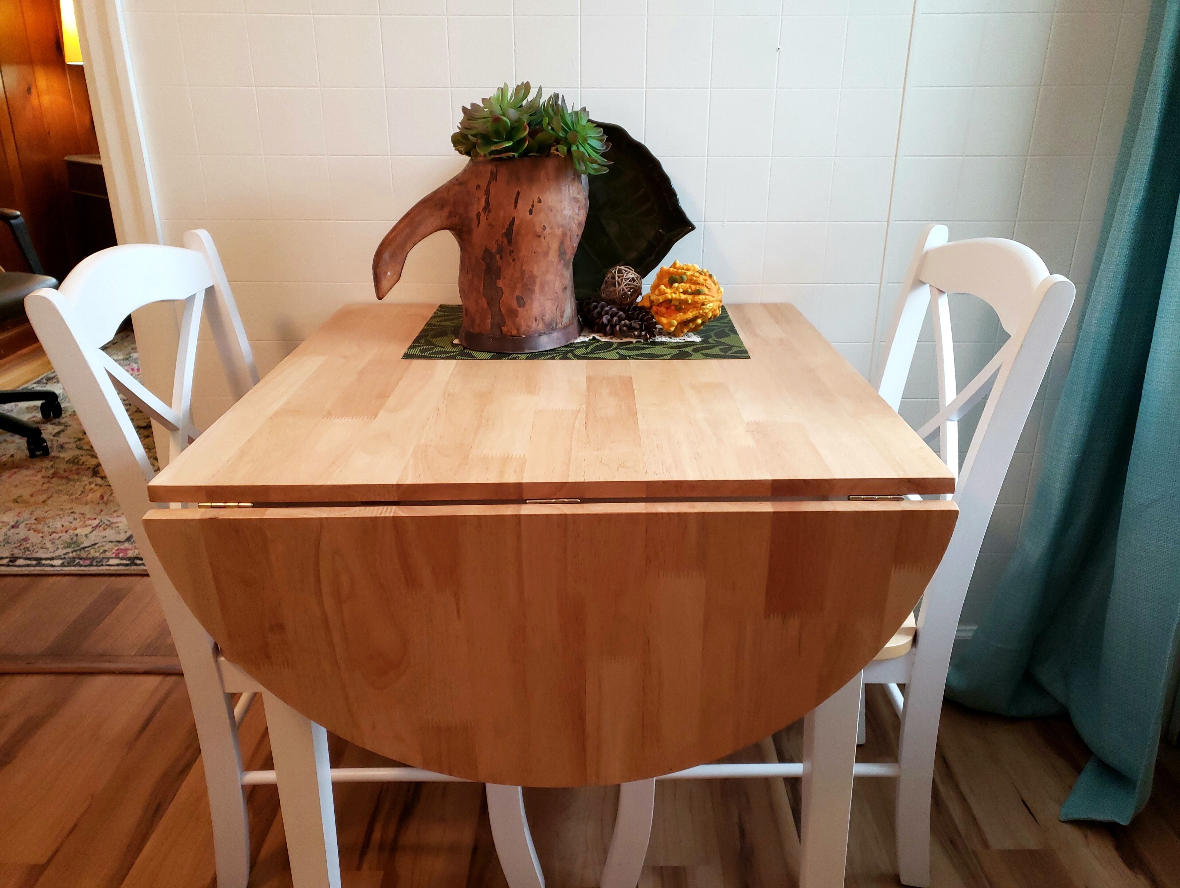 Una mesa redonda de madera con una olla rústica y elementos decorativos, rodeada de sillas blancas.