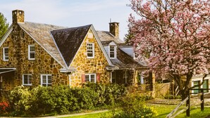 Une maison en pierre avec un toit à forte pente, de nombreuses cheminées et un jardin bien entretenu, agrémenté d’arbres en fleurs.