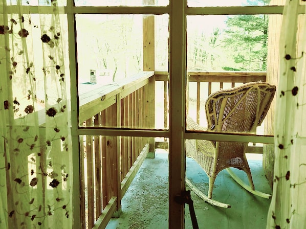 A wooden deck with a wicker rocking chair, a wooden railing, and a view of trees through a window with floral curtains.