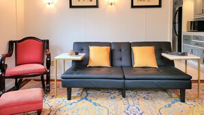 Un salon moderne avec un canapé noir, un fauteuil rouge et une petite table agrémentée d’objets décoratifs. Deux reproductions botaniques encadrées ornent le mur, et on aperçoit une cuisine au fond.
