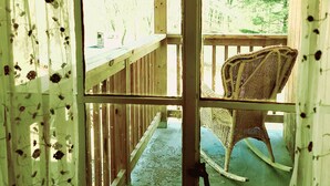 A wooden deck with a wicker rocking chair, a wooden railing, and a view of trees through a window with floral curtains. - ext Stay, 1st fl Luxury Apt, Sanctuary of the Swan Wellness Hotel Swan Rescue (Birdsboro)