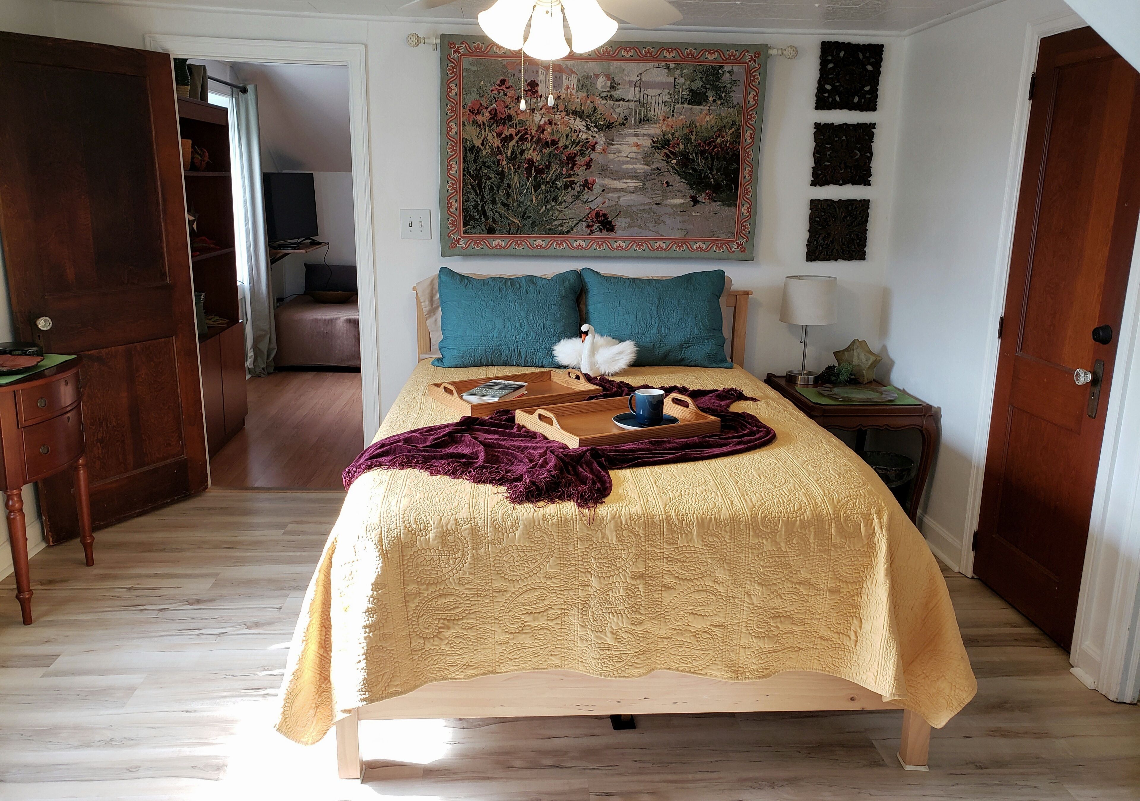 A bedroom with a bed, a wooden dresser, a nightstand, a lamp, a painting, and a door.