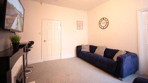 A room with a dark blue sofa, a flat-screen TV mounted on the wall, a white door, and a clock. - Lovely 2-bed Downstairs Apartment (Gateshead)
