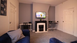 A living room with a fireplace, a television mounted above it, and a blue sofa. - Lovely 2-bed Downstairs Apartment (Gateshead)