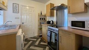 A compact kitchen with a microwave, stove, and sink. There is a wooden cabinet above the stove and a small shelf with kitchen items. - Lovely 2-bed Downstairs Apartment (Gateshead)