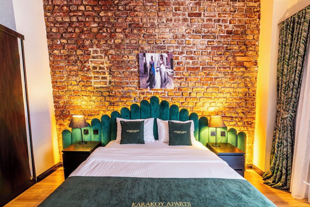 Quarto de hotel com parede de tijolos, cama com cabeceira estofada em verde, duas mesas de cabeceira com abajures e um cobertor verde sobre a cama.