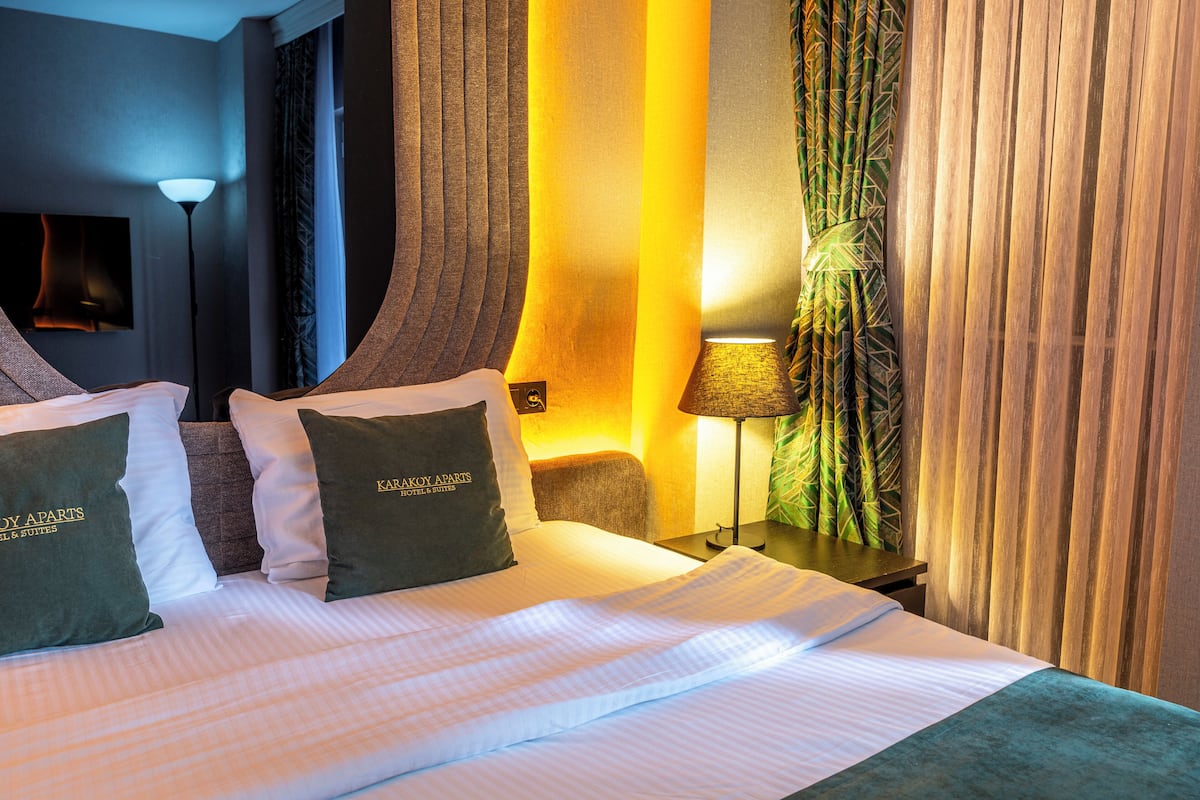 Quarto de hotel com cama, almofadas, mesa de cabeceira, luminária e cortinas.