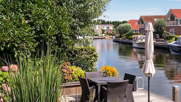 Une terrasse avec une table et des chaises donnant sur un canal où naviguent des bateaux, le tout entouré de verdure et de maisons.