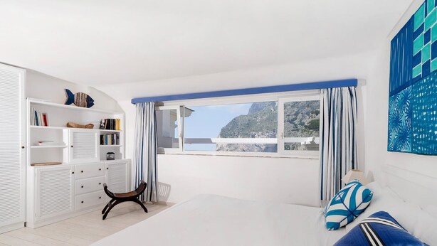 A bedroom with a large bed, a window with curtains, a bookshelf, and a view of the mountains. - Villa Mariuccia Apartment Bea J (Capri)