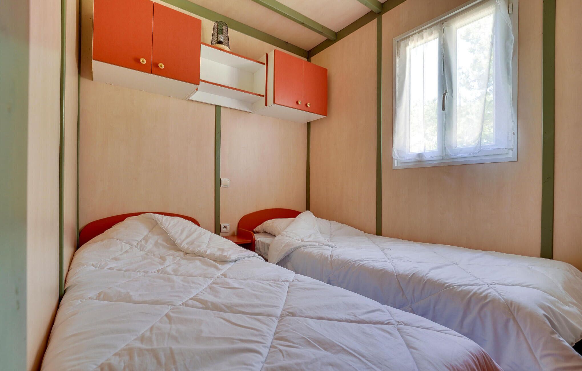 Ein kleiner, kompakter Raum mit zwei Einzelbetten, einem Fenster mit durchscheinenden Vorhängen und einer Küchenzeile mit orangefarbenen Schränken.