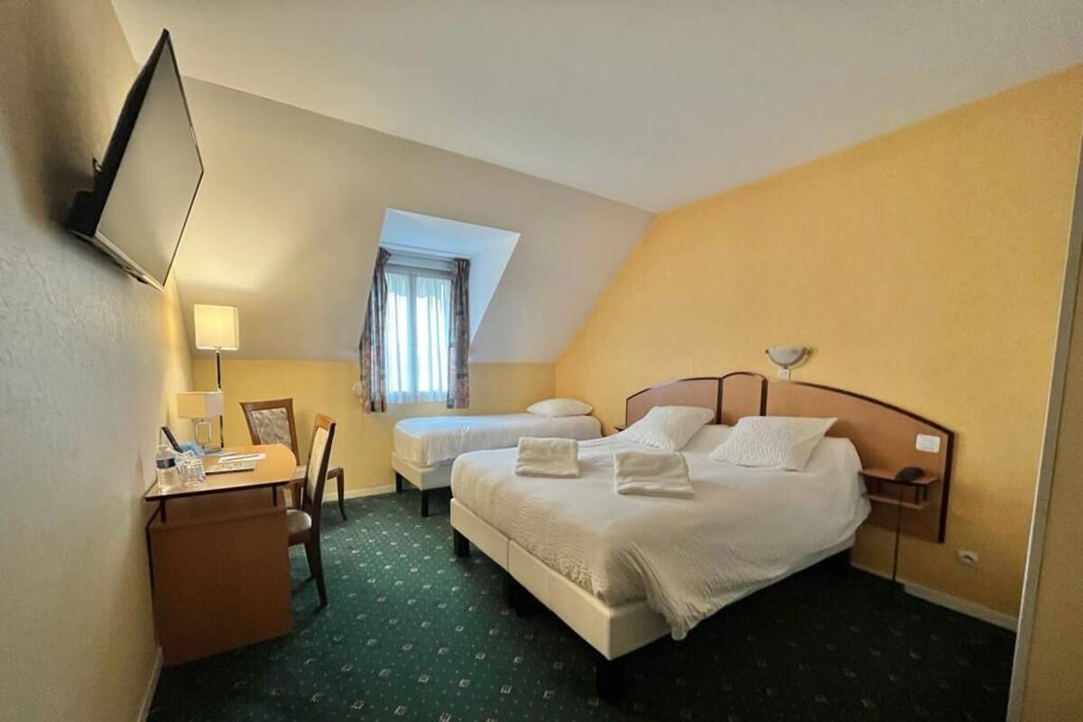 Ein Hotelzimmer mit zwei Betten, einem Schreibtisch mit Stuhl, einem wandmontierten Flachbildfernseher und einem Fenster mit Vorhängen.