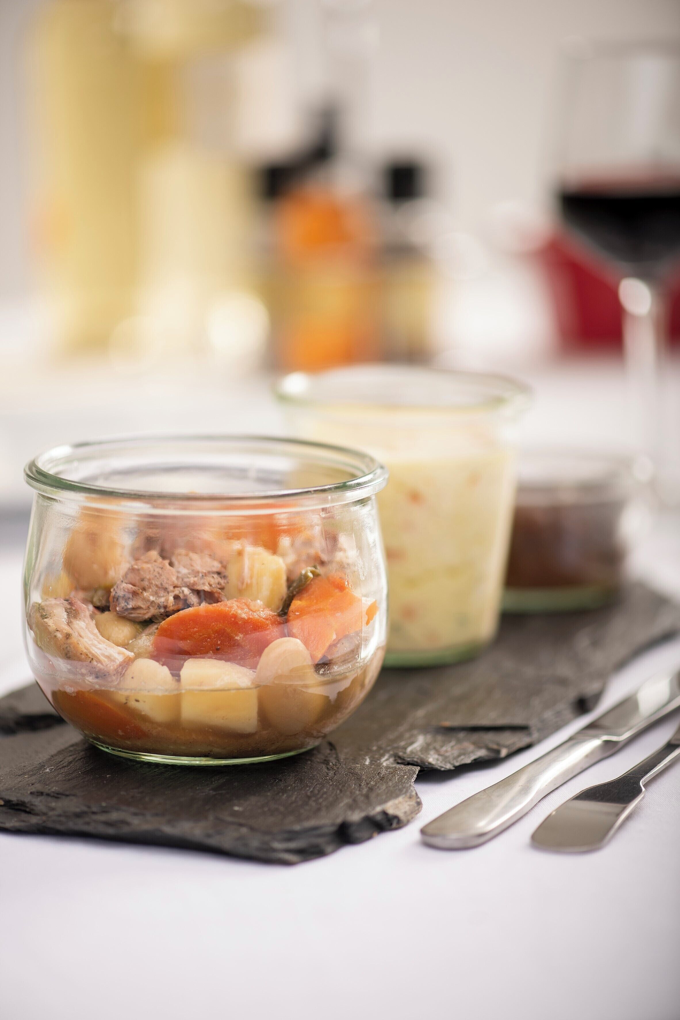 Eine Schüssel mit Eintopf, Gemüse und Fleisch, ein Glas Sahne und ein Gläscher Soße auf einer Schieferplatte.