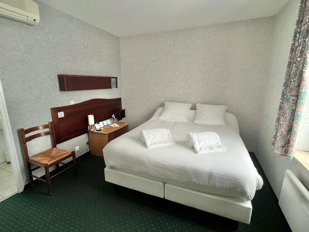 Ein Hotelzimmer mit einem Bett, einem Holzhocker, einem Nachttisch und einer Wand-Klimaanlage.