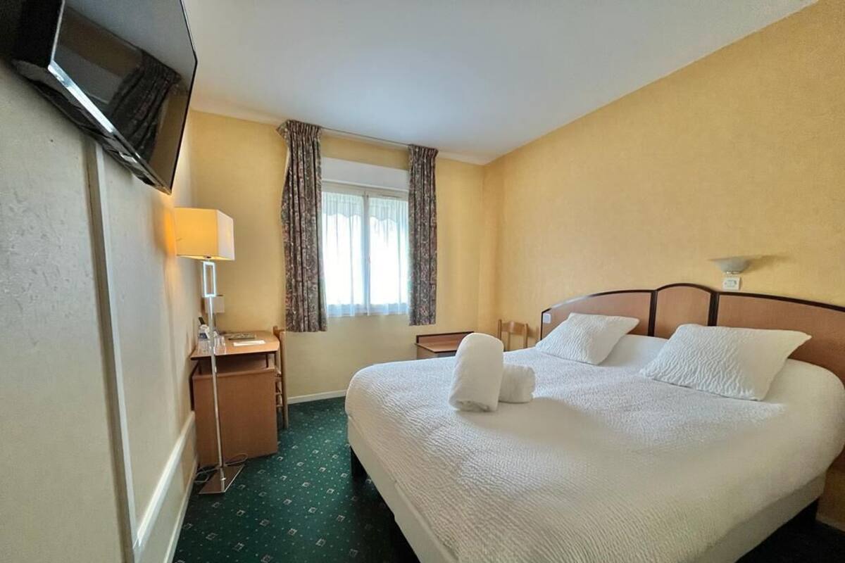 Ein Hotelzimmer mit einem großen Bett, einem Schreibtisch mit Lampe, einem an der Wand befestigten Fernseher und einem Fenster mit Vorhängen.