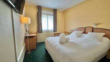 A hotel room with a large bed, a desk with a lamp, a television mounted on the wall, and a window with curtains.