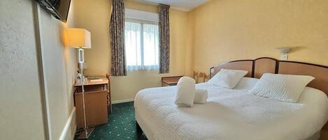 A hotel room with a large bed, a desk with a lamp, a television mounted on the wall, and a window with curtains.