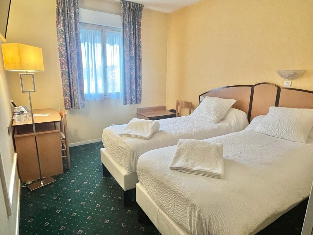 Ein Hotelzimmer mit zwei Betten, einem Schreibtisch, einer Lampe und einem Fenster mit Vorhängen.