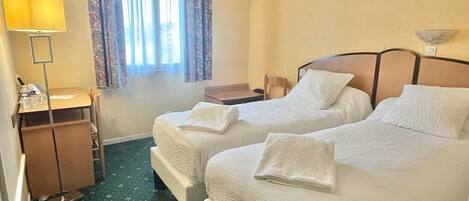 A hotel room with two beds, a desk, a lamp, and a window with curtains.