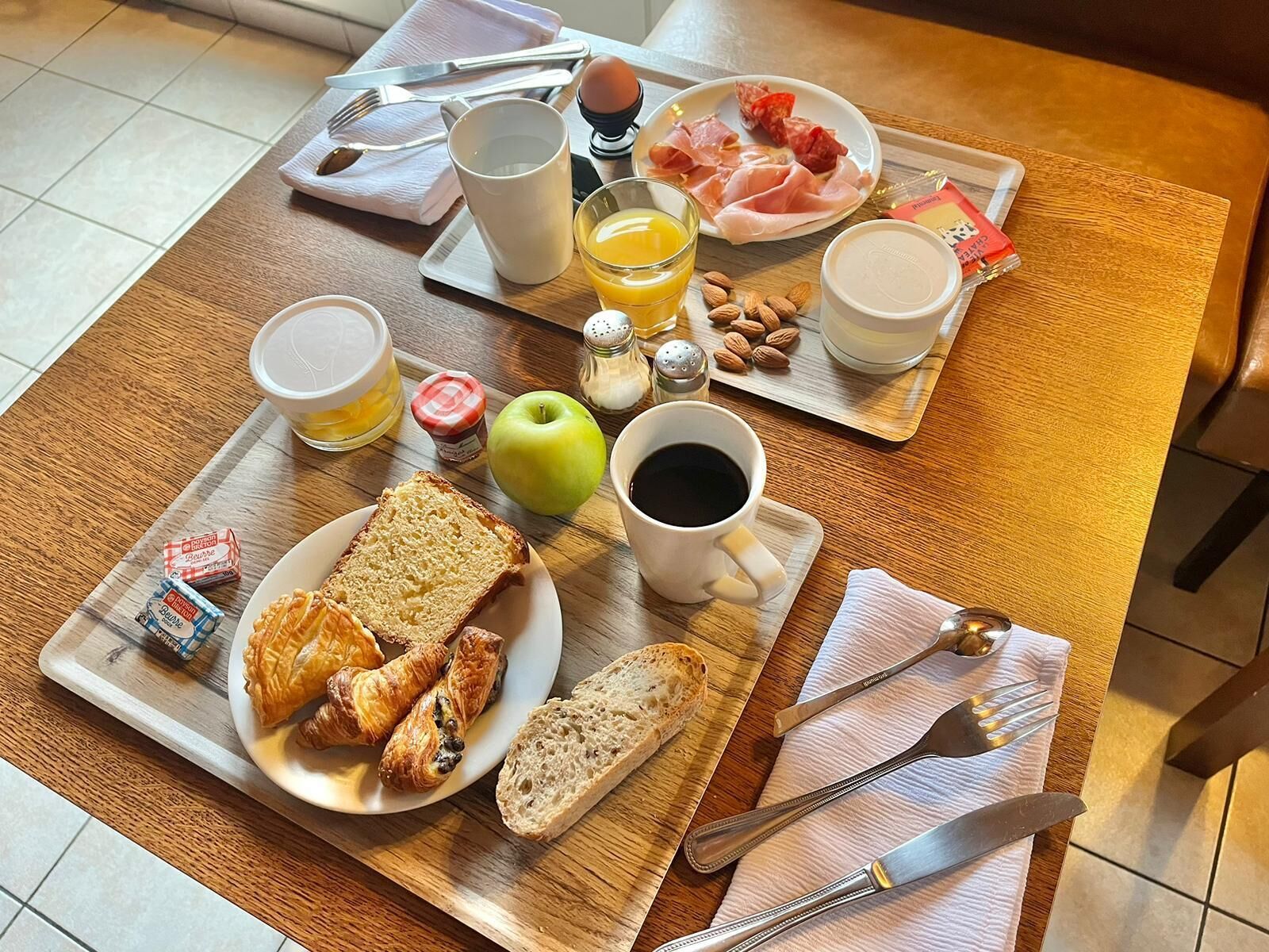 Bandeja de desayuno con cruasanes, pan, una manzana, un vaso de jugo de naranja, un plato de jamón, una taza de café y aderezos.