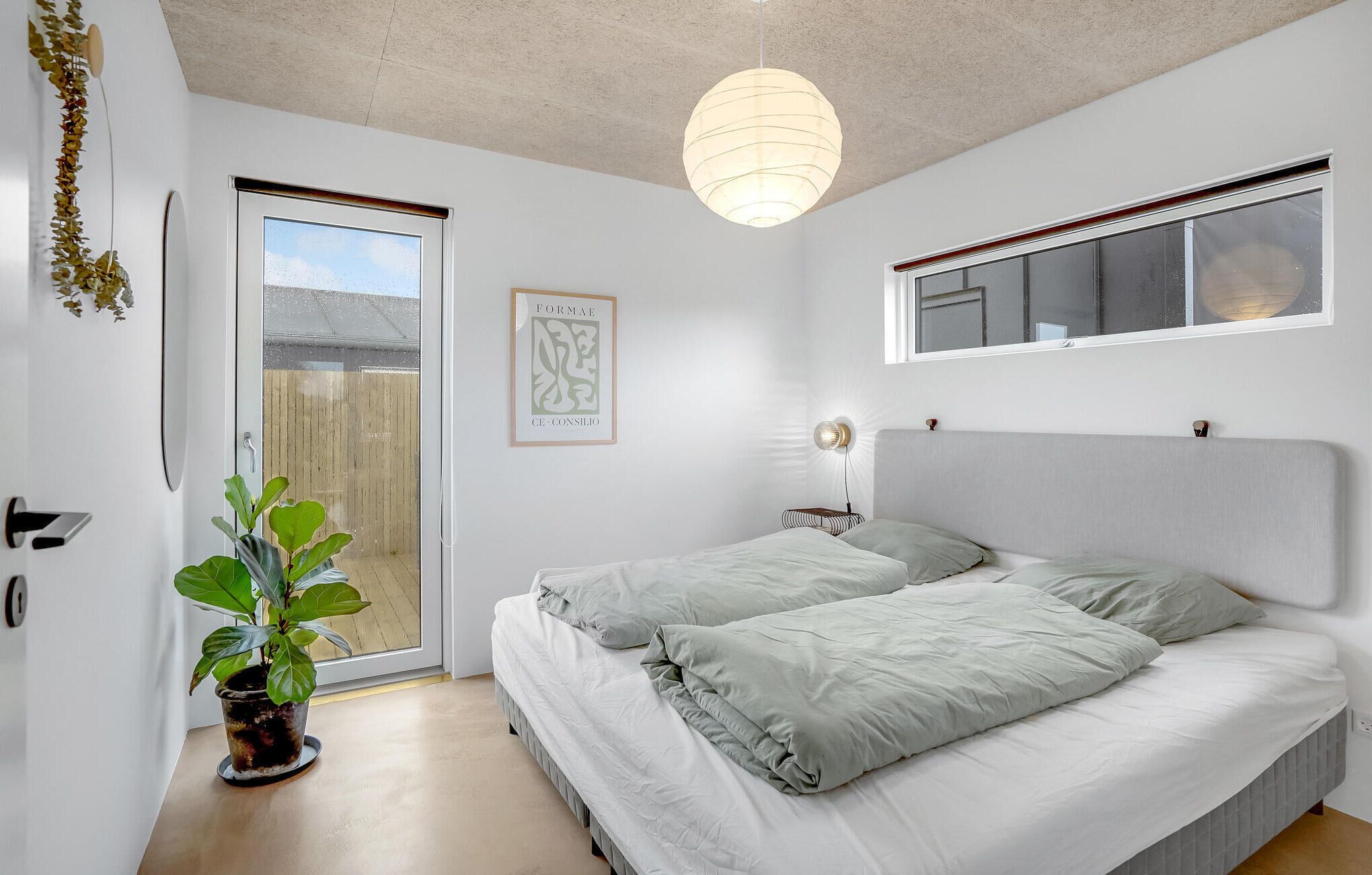 A bedroom with a bed, a bedside table, a wall-mounted lamp, a window, and a door leading to a balcony.