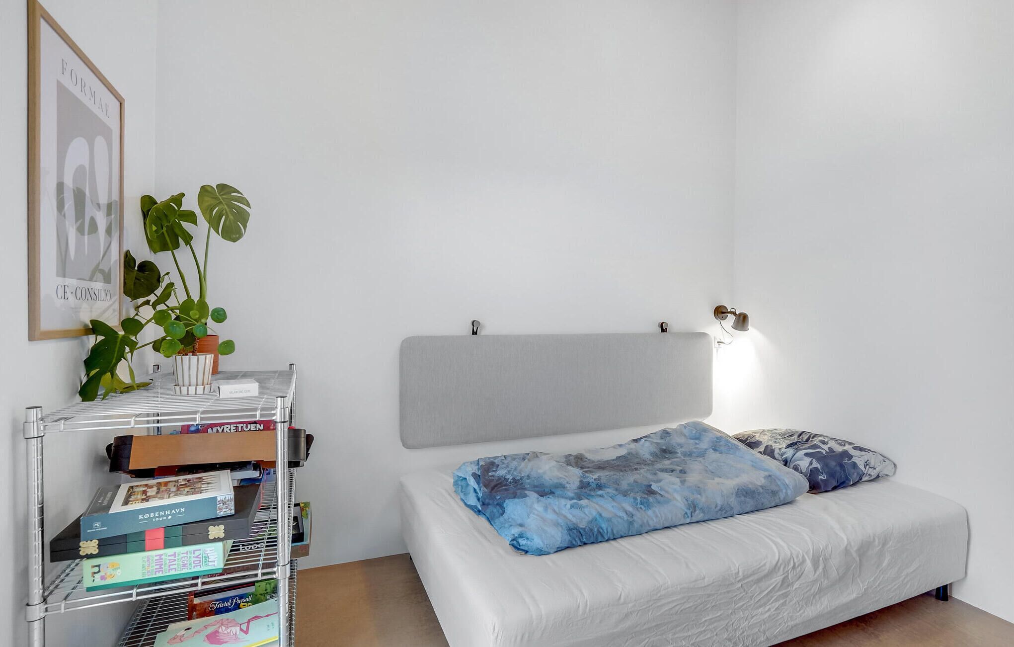 A minimalist bedroom with a bed, a nightstand, a wall-mounted shelf with books, and a potted plant.