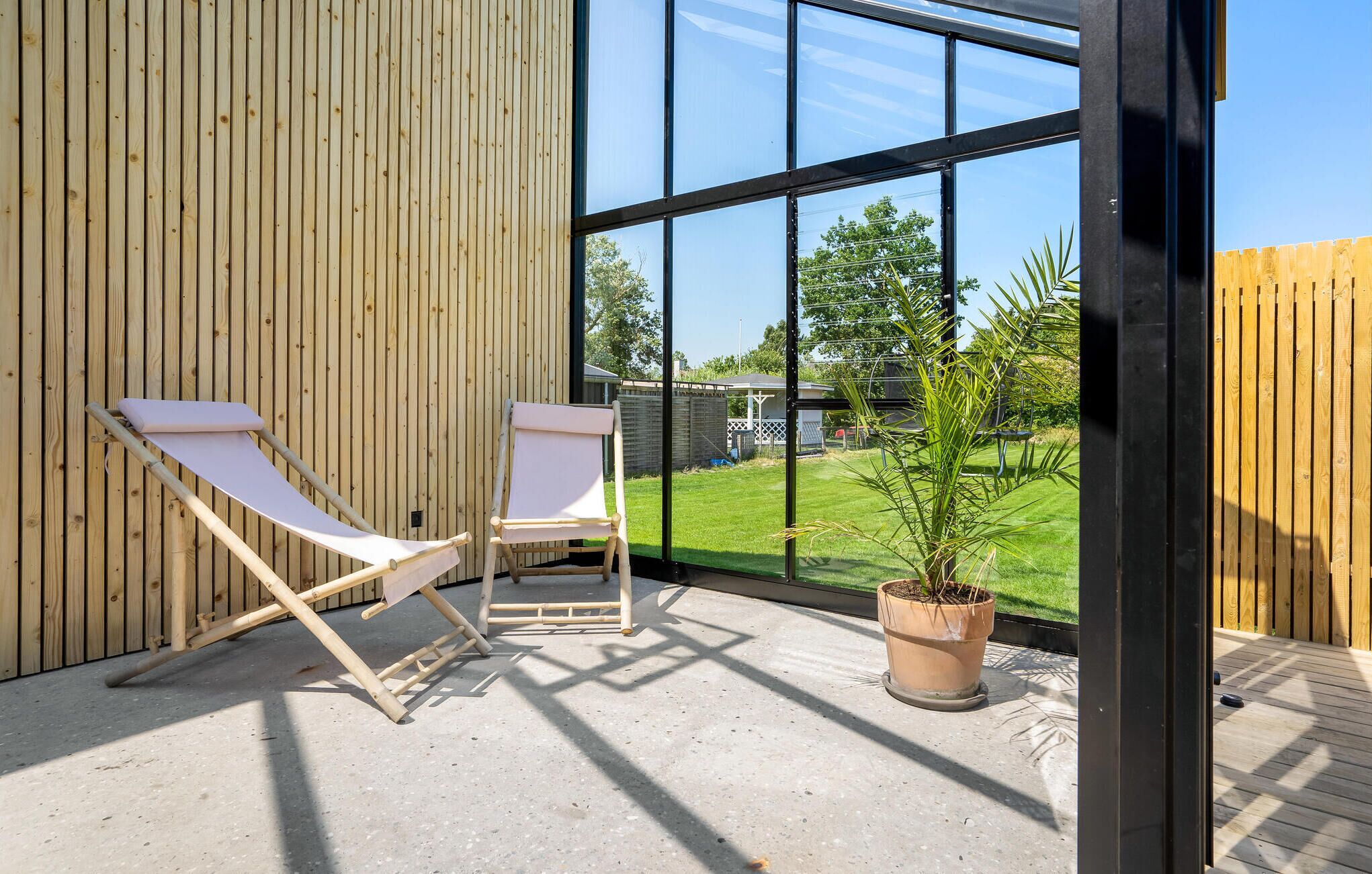 A covered patio area with two white lounge chairs, a potted plant, and a view of a green lawn and trees through large windows.