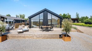 A modern house with a large glass front, a wooden deck with outdoor furniture, and a gravel driveway. - 4 bedroom cozy home in Haderslev (Haderslev)