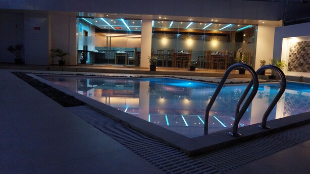 A swimming pool with a glass barrier, a poolside area with tables and chairs, and a building with a modern design. - Hotel Sea Port (Port Blair)