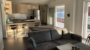 A modern kitchen with wooden cabinets, a dining area with a grey sofa and a table, and a view of the sea through a sliding door.