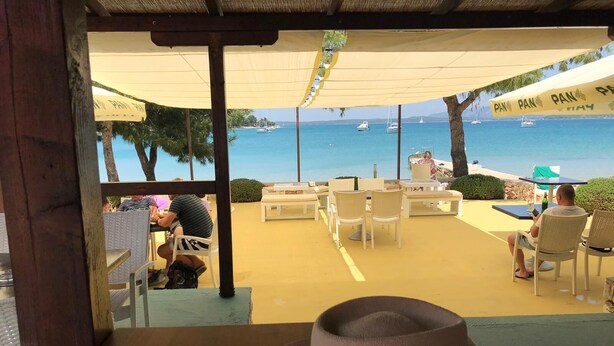 An outdoor seating area with tables and chairs, overlooking a beach with boats.