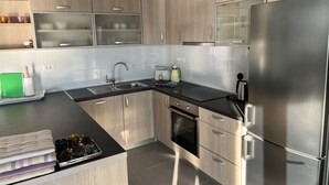 A modern kitchen with light wood cabinets, a stainless steel refrigerator, a built-in oven, and a black countertop.