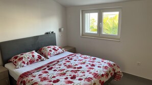 A bedroom with a bed, nightstands, a window with a view of greenery, and a wall-mounted light.