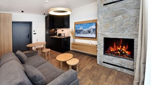 A modern living room with a grey sofa, wooden tables, a stone fireplace, and a kitchen area with dark cabinets and a pendant light.