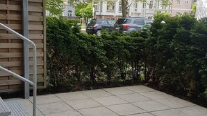 Pátio pavimentado com escadaria de metal, sebes verdes densas e carros estacionados ao fundo.