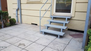 Escadas metálicas externas levam a uma entrada de edifício, com uma porta e janela visíveis.
