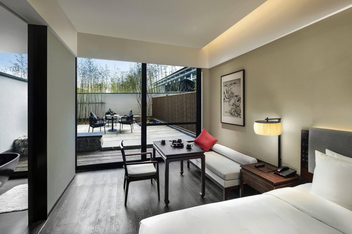 Habitación de hotel con cama, escritorio con silla, sofá, mesita de noche con lámpara y vistas al exterior.