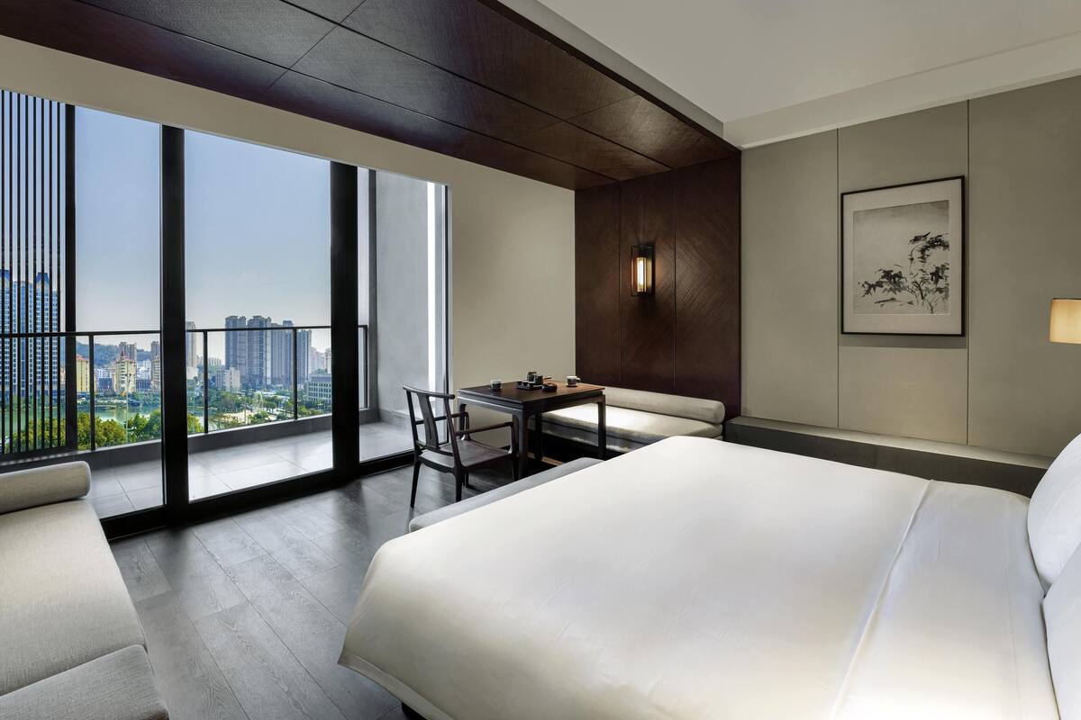 Un cuarto de hotel con una cama grande, una mesa de comedor con sillas, un sofá, vistas de la ciudad a través de amplias ventanas y una obra de arte enmarcada en la pared.