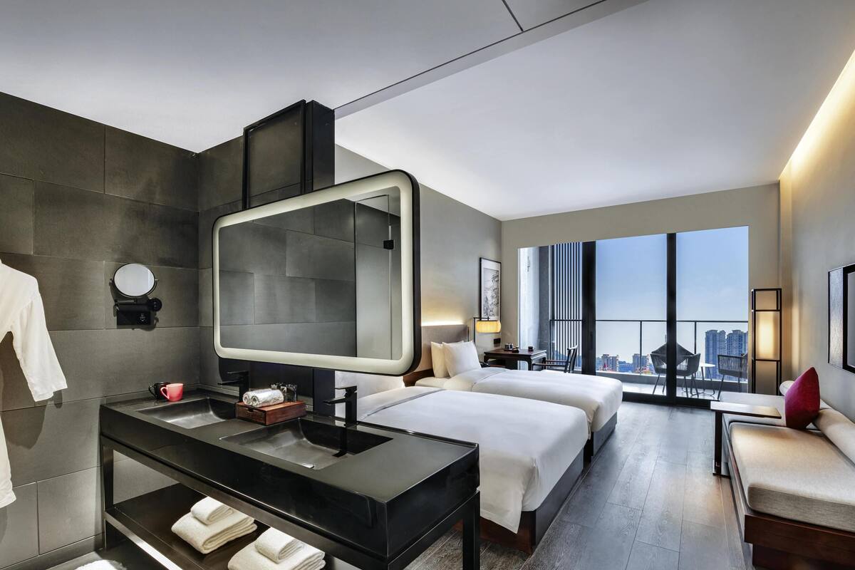 Una habitación de hotel moderna con un espejo grande, dos camas, una tocador con lavamanos y vistas a la ciudad.