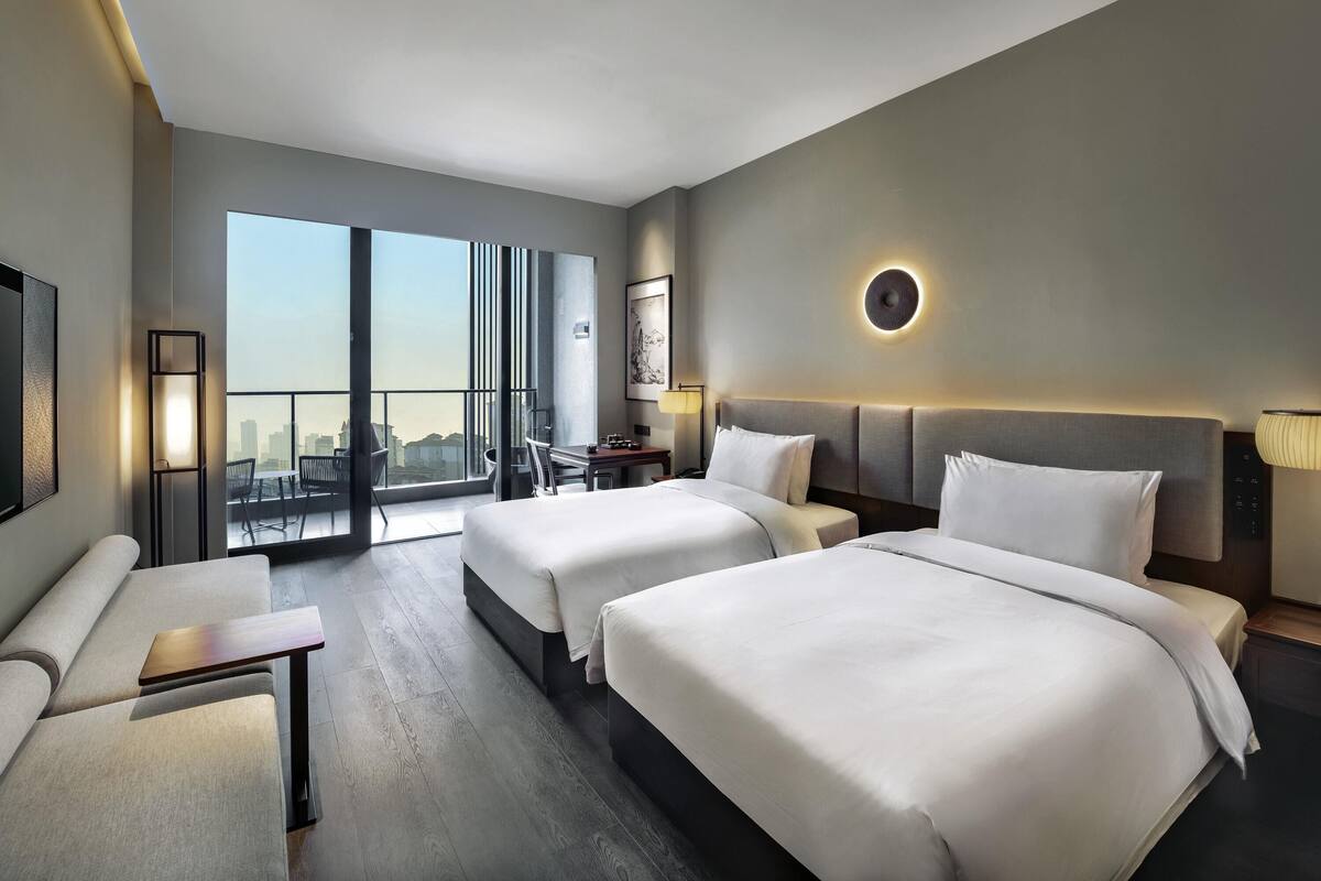 Una habitación de hotel moderna con dos camas, un escritorio, una silla y un amplio ventanal con vistas a la ciudad.