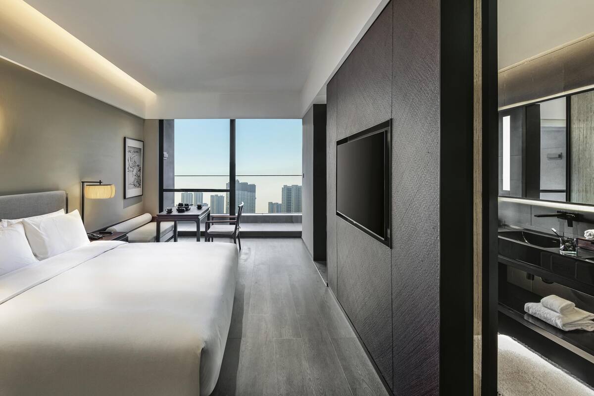 Una habitación de hotel moderna con una cama grande, un escritorio con silla, un televisor de pantalla plana, un espejo amplio y vistas a la ciudad.