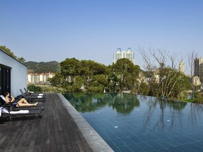 Piscina infinita en la azotea con sillones de descanso, una terraza de madera y vistas a edificios y árboles de la ciudad.