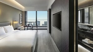 Habitación de hotel moderna con una cama grande, un escritorio, un televisor de pantalla plana en la pared y un amplio ventanal con vistas a la ciudad.