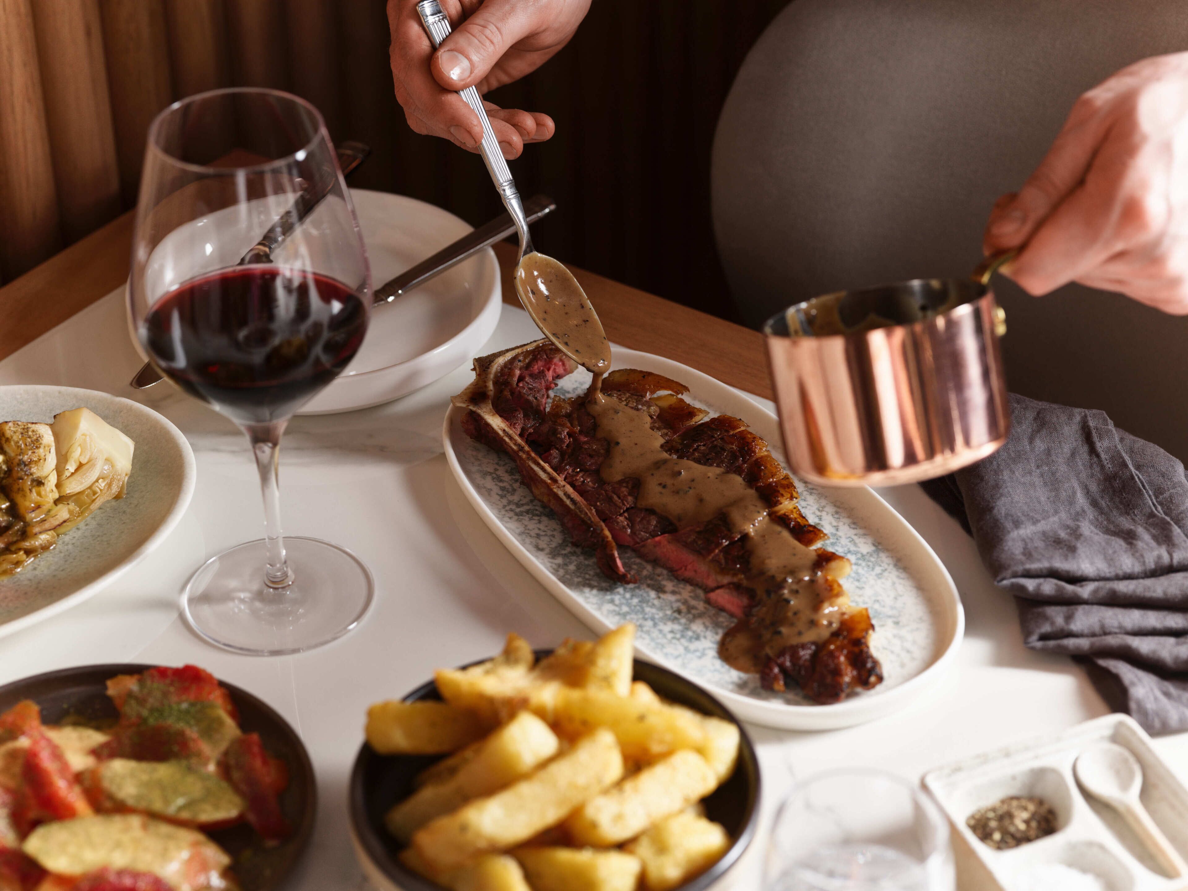 A dining table with a steak, a glass of red wine, and a plate of fries.