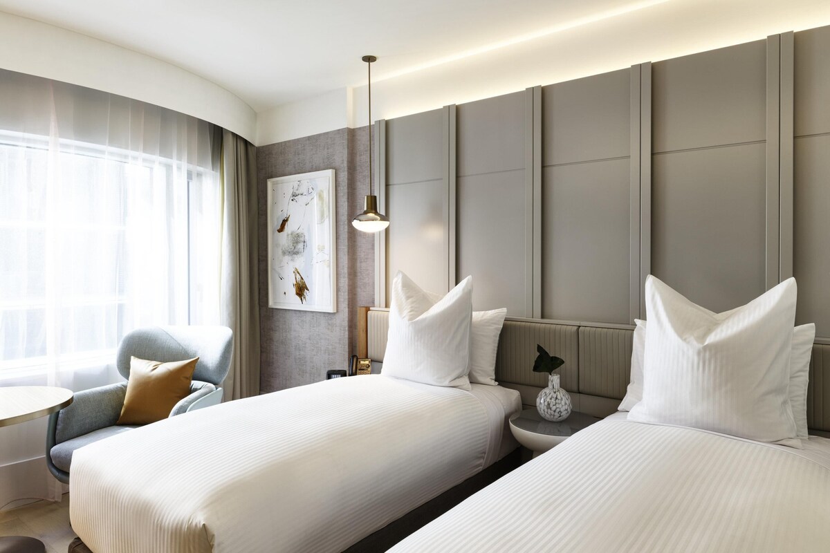 Une chambre d’hôtel avec deux lits, un coin salon comprenant un fauteuil, une table et une grande fenêtre avec des rideaux.
