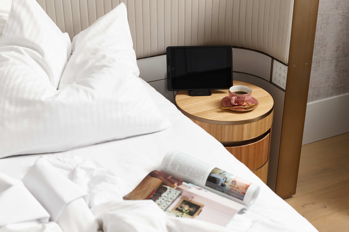 Un lit bien fait, avec du linge de lit blanc, une tablette posée sur une table de chevet en bois, une tasse de café et un magazine sur le lit.