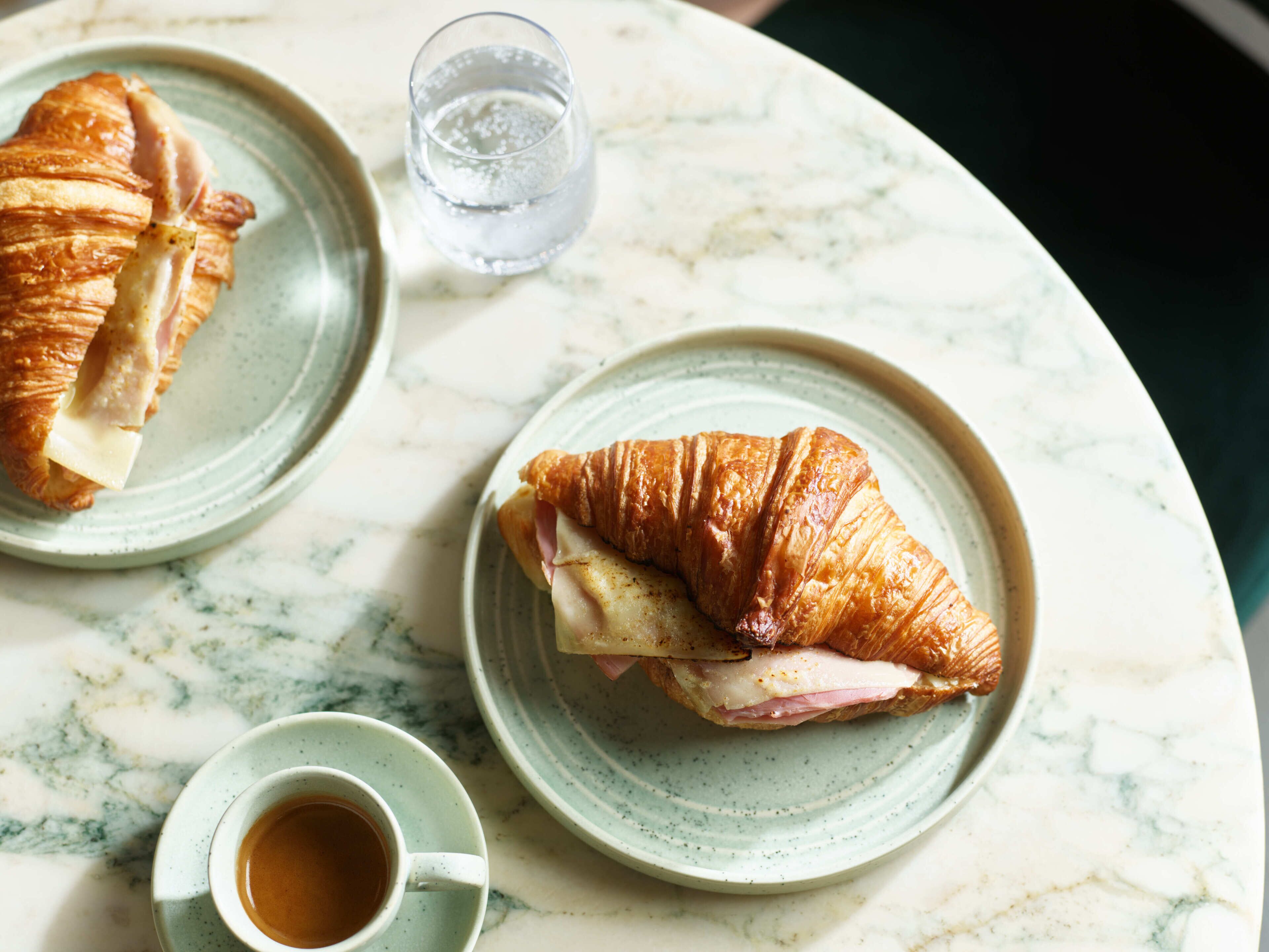 Dois croissants em pratos com uma chávena de café e um copo de água sobre uma mesa de mármore.