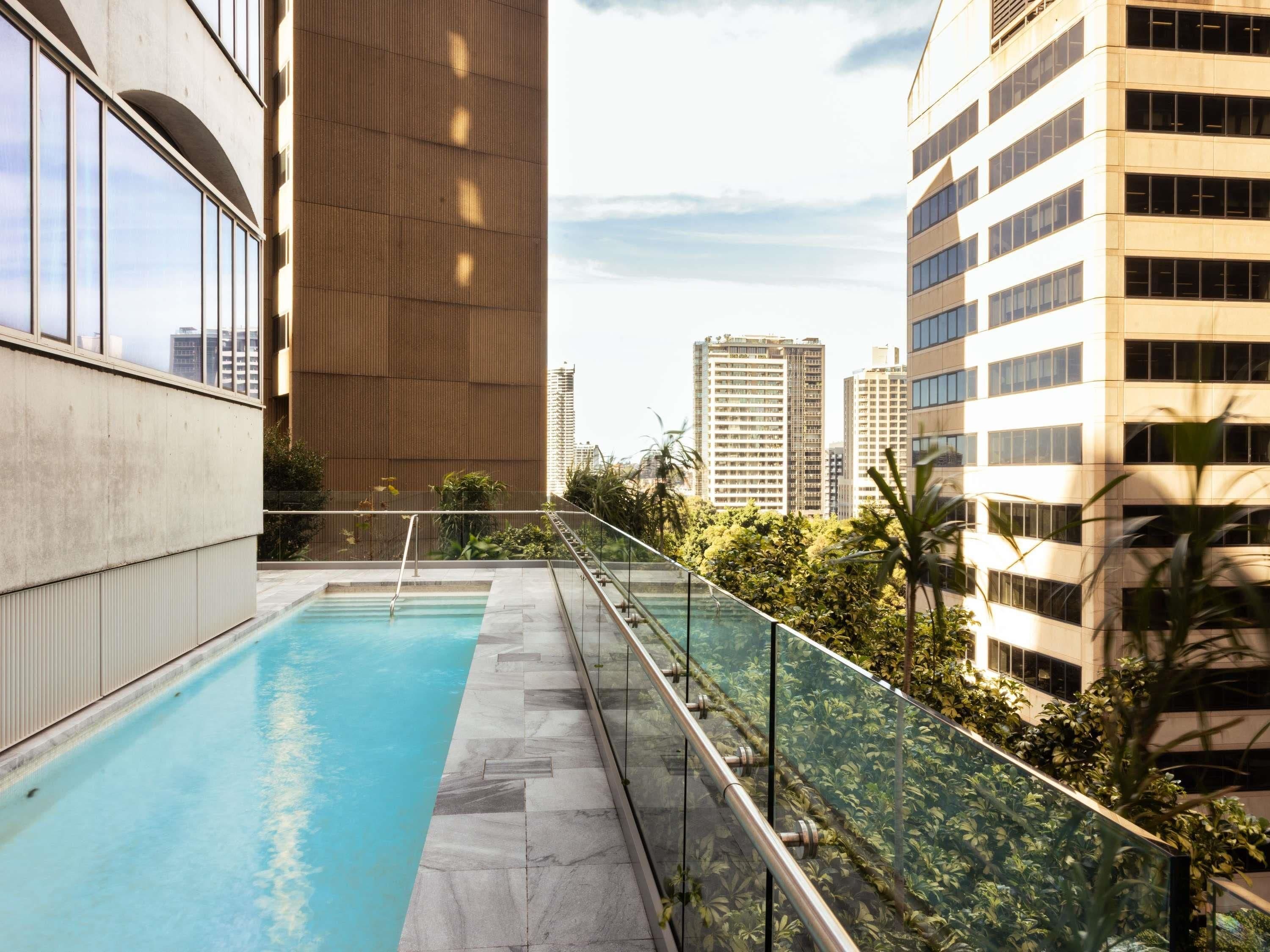 Una piscina en la azotea con barandilla de cristal, rodeada de follaje y vistas a un paisaje urbano.